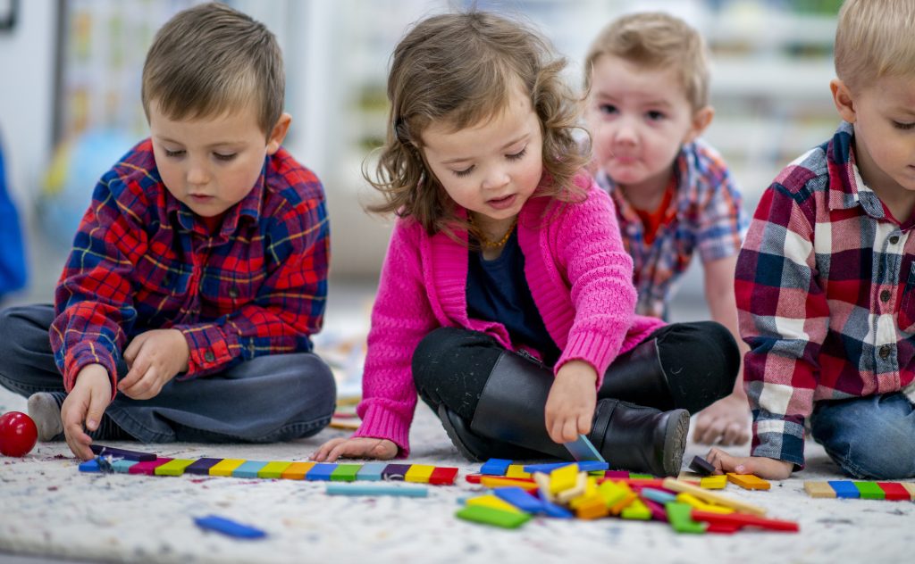 A sweet little blond haired girl sits on the floor of her preschool classroom as she plays with colorful blocks. She is dressed casually and is focused on the activity as her peers play in the background. - preschool franchise