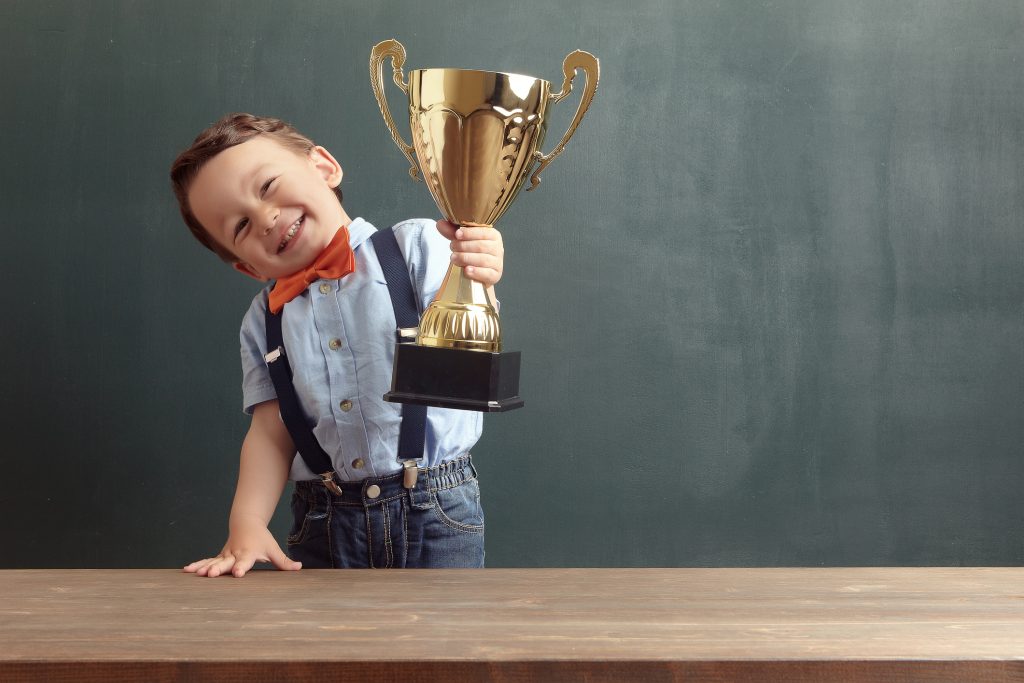 A cute, smiling 2-3 years old boy is standing behind a wooden table and raising a golden trophy with his hand. Little boy is wearing an orange bow tie and blue trousers with suspenders. - 